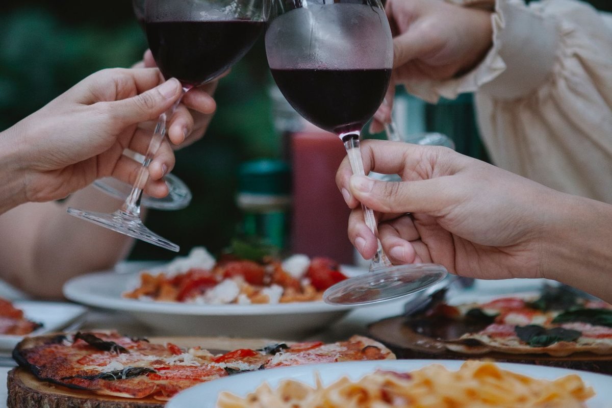 Duas pessoas brindando com taças de vinho tinto sobre uma mesa com pizza e massa