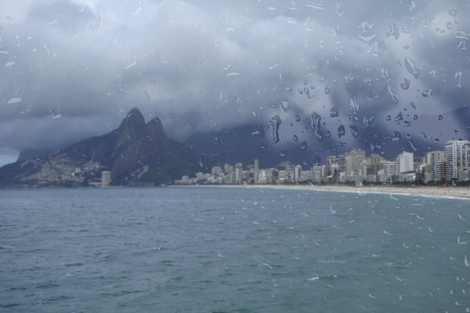 Morro Dois Irmãos e a praia de Ipanema vistos através de uma janela molhada de chuva, em dia frio e nublado no Rio de Janeiro