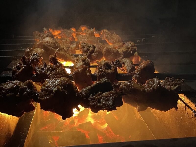 Espetinhos de carne assando sobre brasa acesa em churrasqueira à noite, com fumaça subindo — que vinho combina com churrasco muda conforme o que está na grelha
