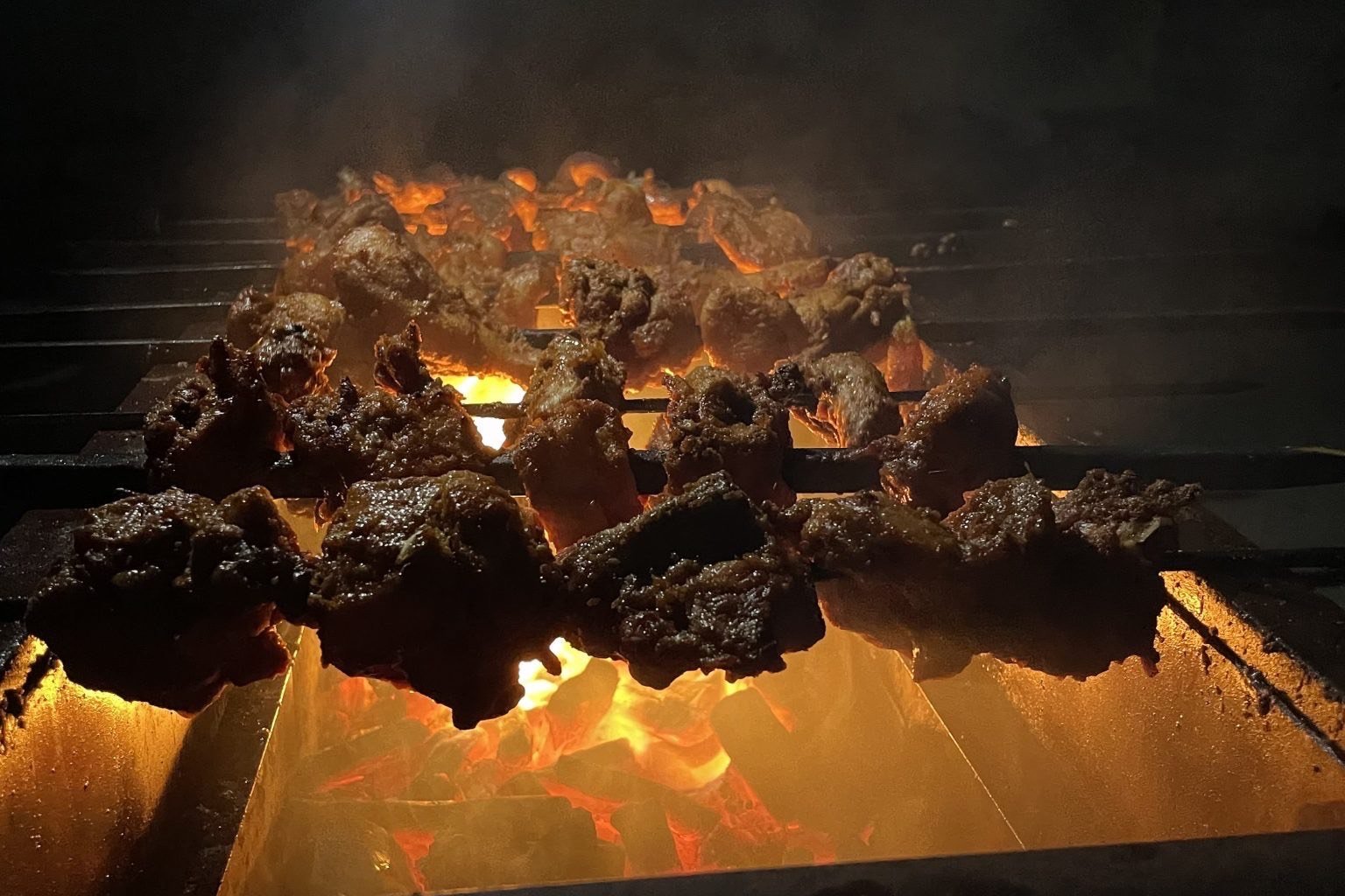 Espetinhos de carne assando sobre brasa acesa em churrasqueira à noite, com fumaça subindo — que vinho combina com churrasco muda conforme o que está na grelha