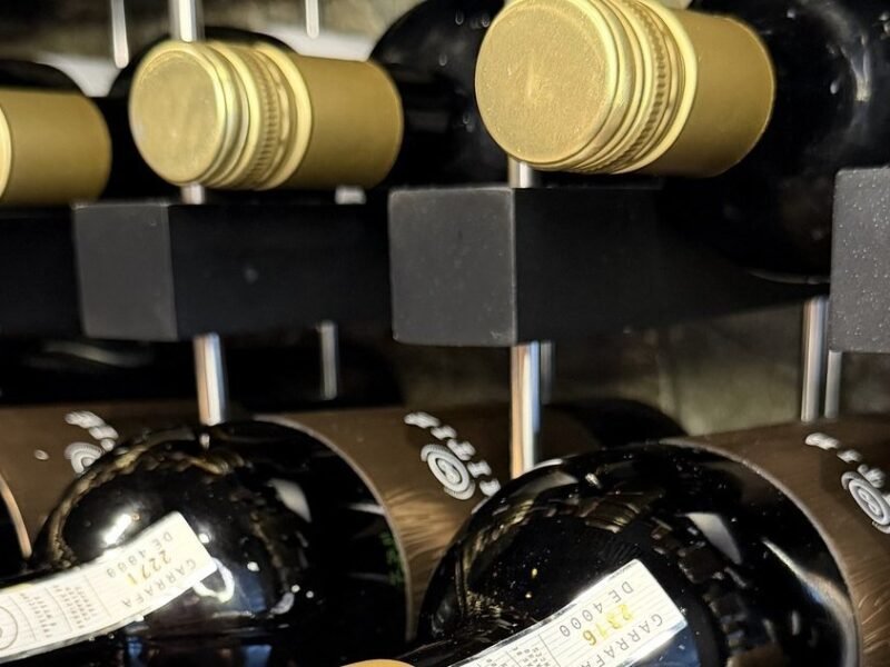 Garrafas de vinho deitadas na prateleira da adega da Better Wine, com cápsulas douradas