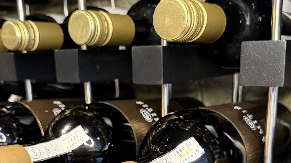 Garrafas de vinho deitadas na prateleira da adega da Better Wine, com cápsulas douradas