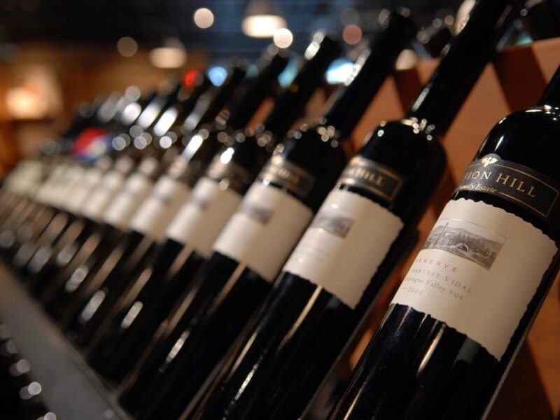 Fileira de garrafas de vinho tinto deitadas em prateleira de madeira de adega, com luz baixa ao fundo