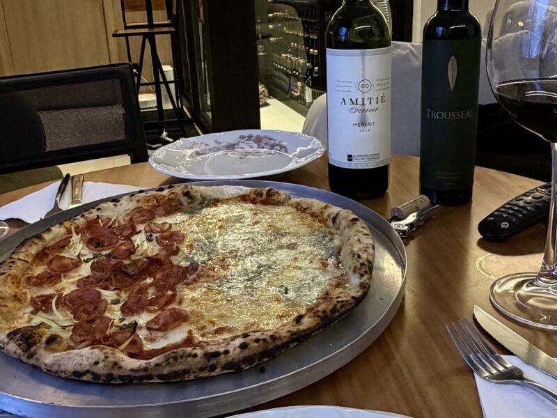 Pizza meio calabresa meio quatro queijos numa mesa posta em casa, com uma garrafa de Amitié Terroir Merlot e a adega da Better Wine acesa ao fundo