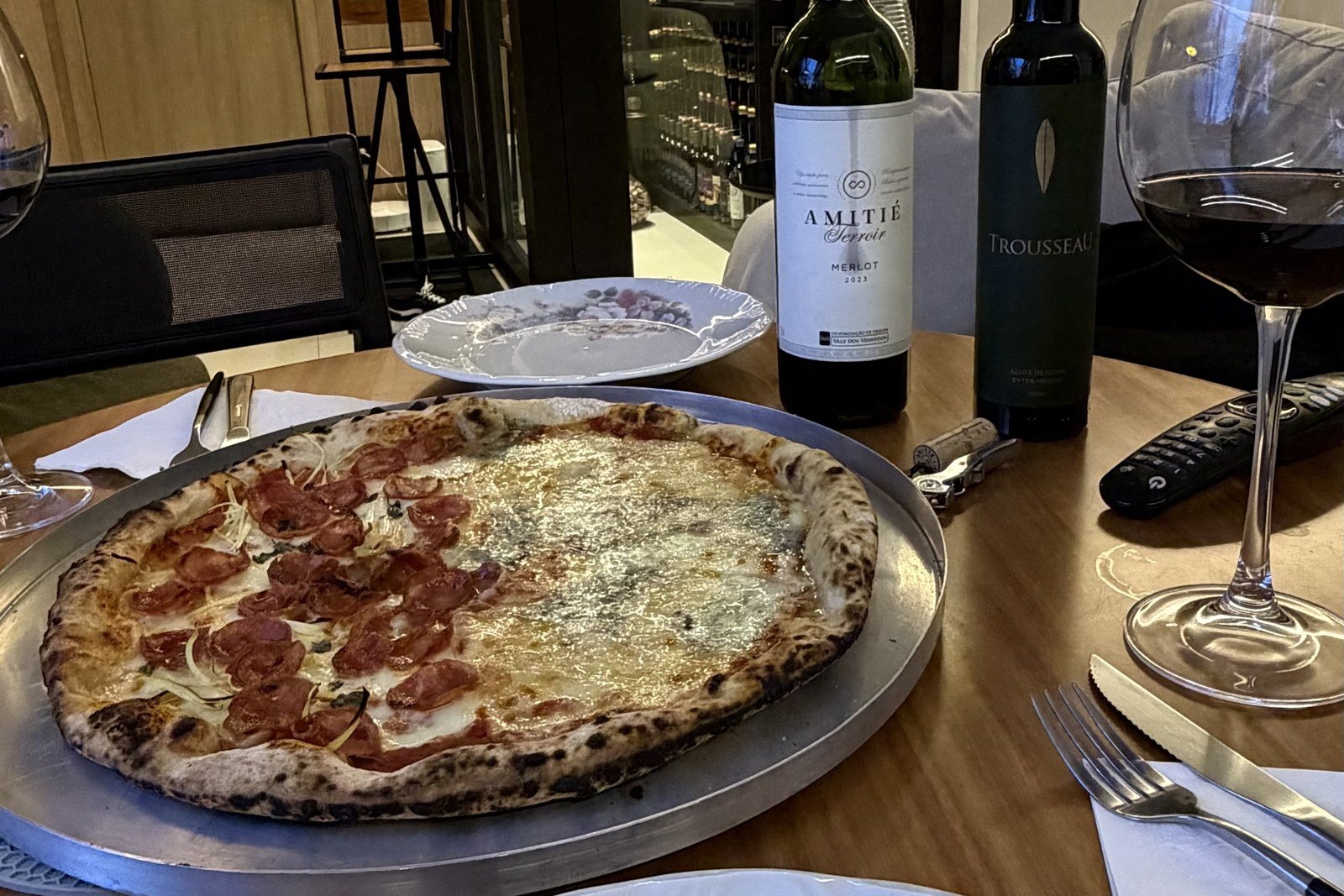 Pizza meio calabresa meio quatro queijos numa mesa posta em casa, com uma garrafa de Amitié Terroir Merlot e a adega da Better Wine acesa ao fundo