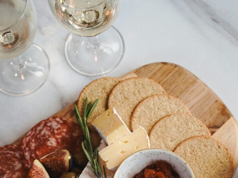 Tábua de frios e queijos com salame, brie, figos, azeitonas e torradas ao lado de duas taças de vinho branco