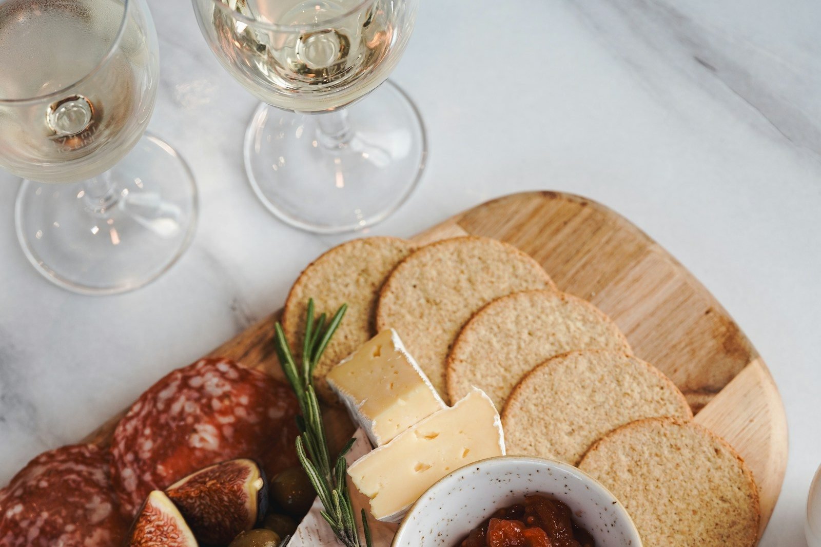 Tábua de frios e queijos com salame, brie, figos, azeitonas e torradas ao lado de duas taças de vinho branco