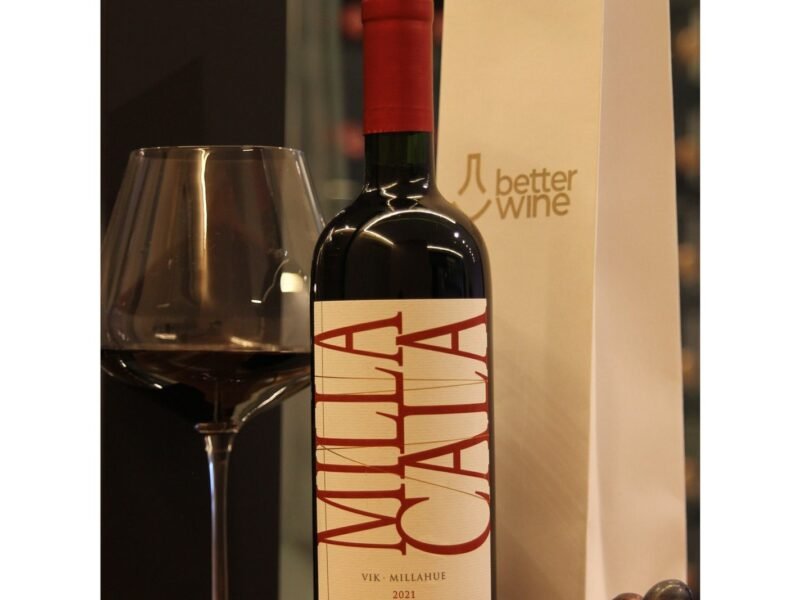 Taça de vinho vazia, com bojo largo e haste longa, ao lado de uma garrafa de tinto Milla Cala, com o logo da Better Wine ao fundo