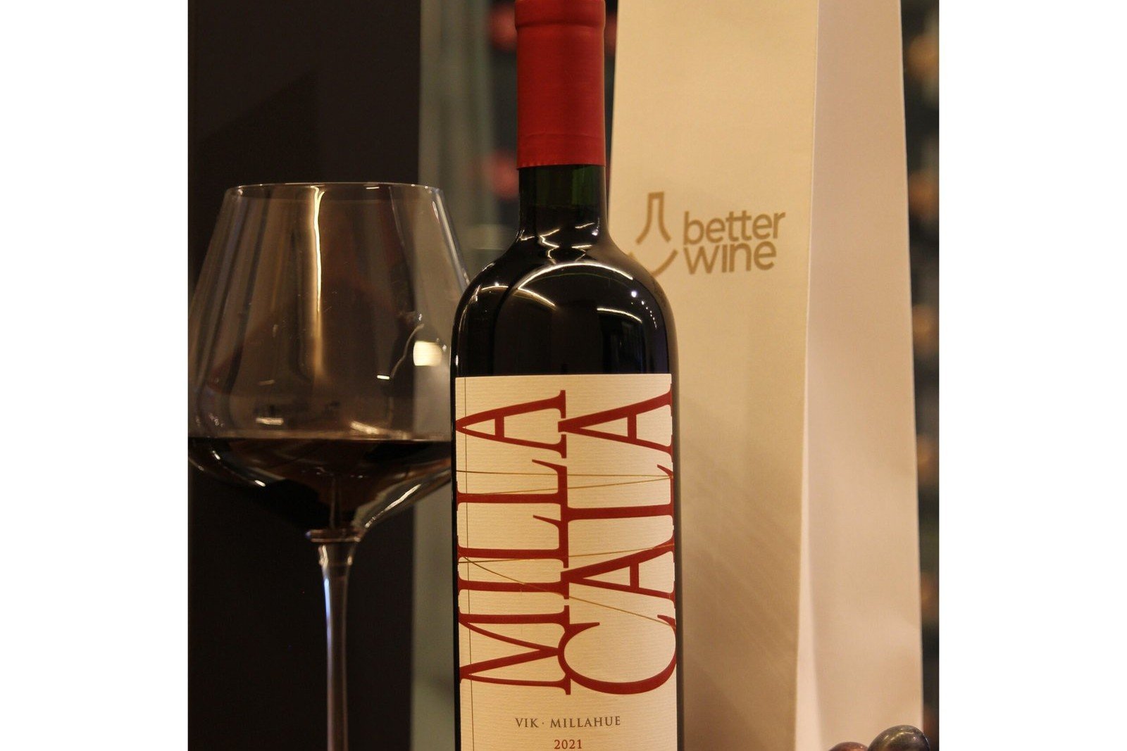 Taça de vinho vazia, com bojo largo e haste longa, ao lado de uma garrafa de tinto Milla Cala, com o logo da Better Wine ao fundo