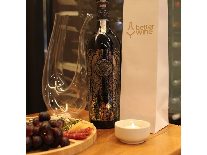 Garrafa de Patrimony Black Label ao lado de um decanter, taça, uvas e uma vela acesa, com o logo da Better Wine ao fundo