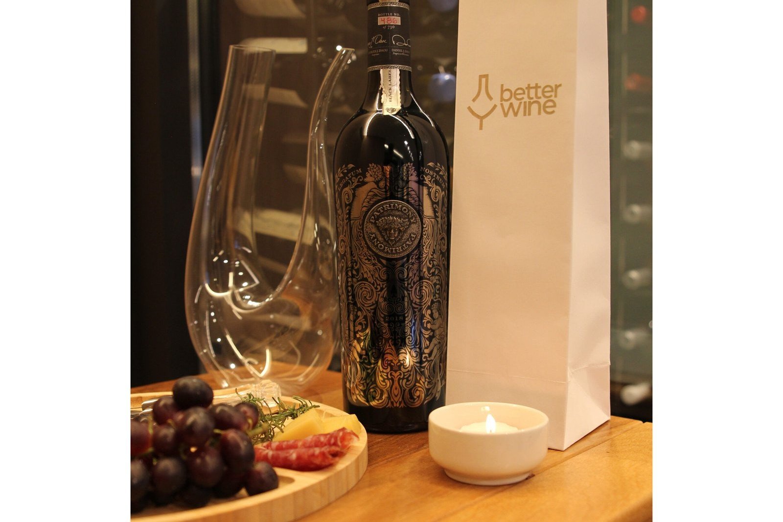 Garrafa de Patrimony Black Label ao lado de um decanter, taça, uvas e uma vela acesa, com o logo da Better Wine ao fundo