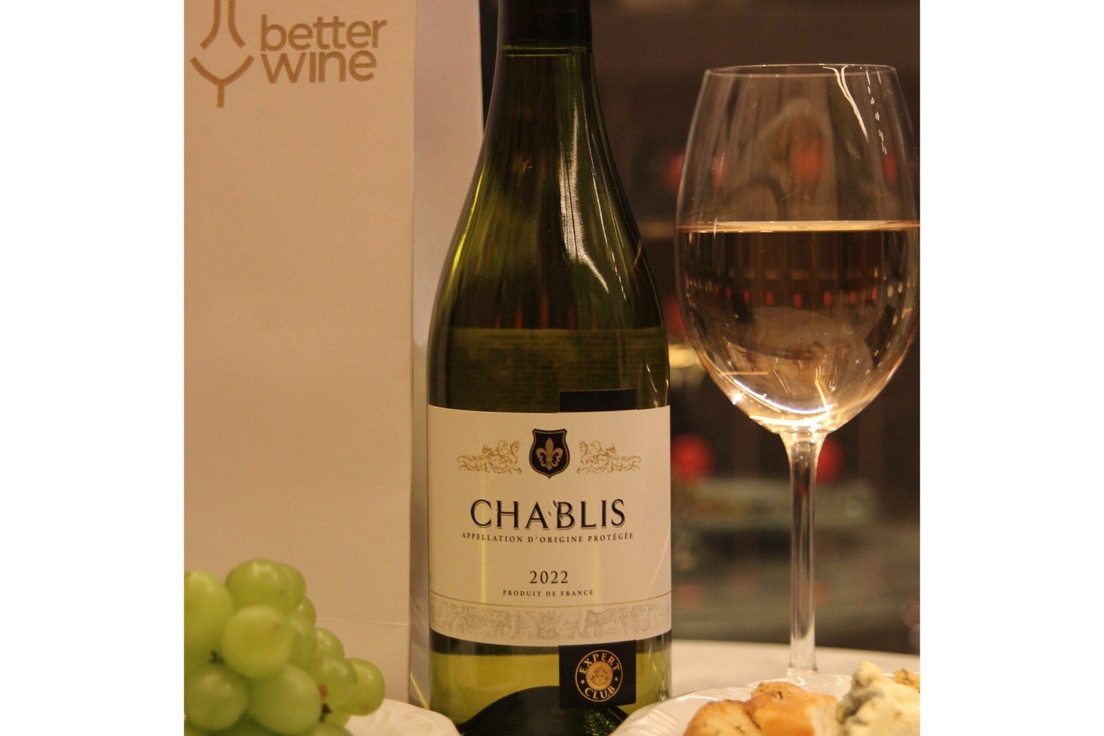 Garrafa de Chablis 2022 ao lado de uma taça de vinho branco, uvas verdes e queijo, com o logo da Better Wine ao fundo